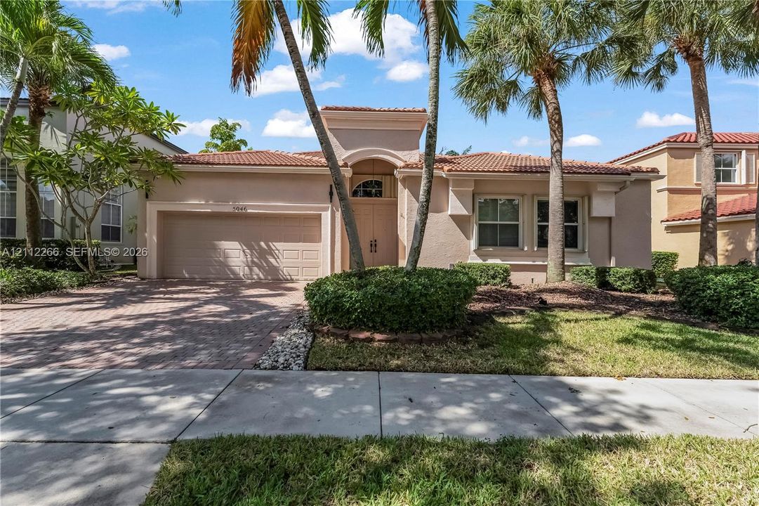 Single-story residence featuring a tile roof, stucco exterior, arched entryway, brick-paver driveway, and mature palm trees