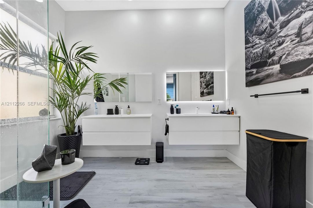Dual white floating vanities featuring integrated sinks and minimalist cabinetry, paired with rectangular backlit mirrors, glass-enclosed shower, and wood-finish flooring