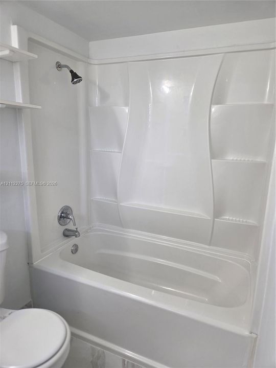 White acrylic bathtub and shower surround featuring integrated shelving, chrome fixtures, and a wall-mounted shower head