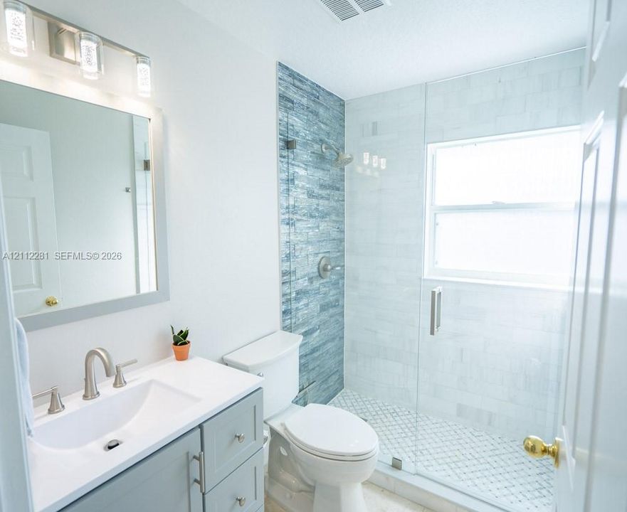 Bathroom featuring a vanity with a white countertop and rectangular undermount sink, a walk-in shower with glass enclosure, textured blue tile accent wall, and three-light brushed-nickel fixture above the mirror