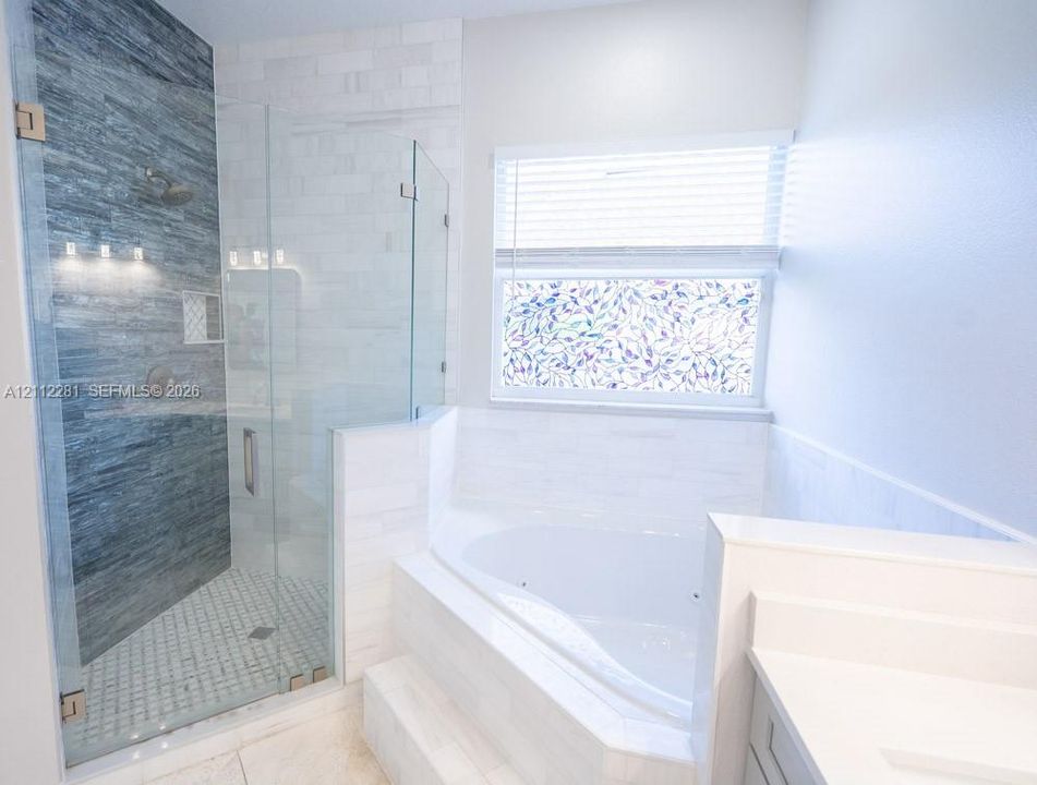 Walk-in glass-enclosed shower with horizontal textured tiling and built-in shelving, alongside a white jetted soaking tub with tile surround