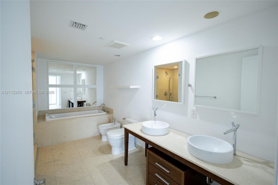 Double vanity featuring two oval vessel sinks, stone-finish countertop, dark wood cabinetry, and wall-mounted mirrors, complemented by a drop-in soaking tub with a tiled surround