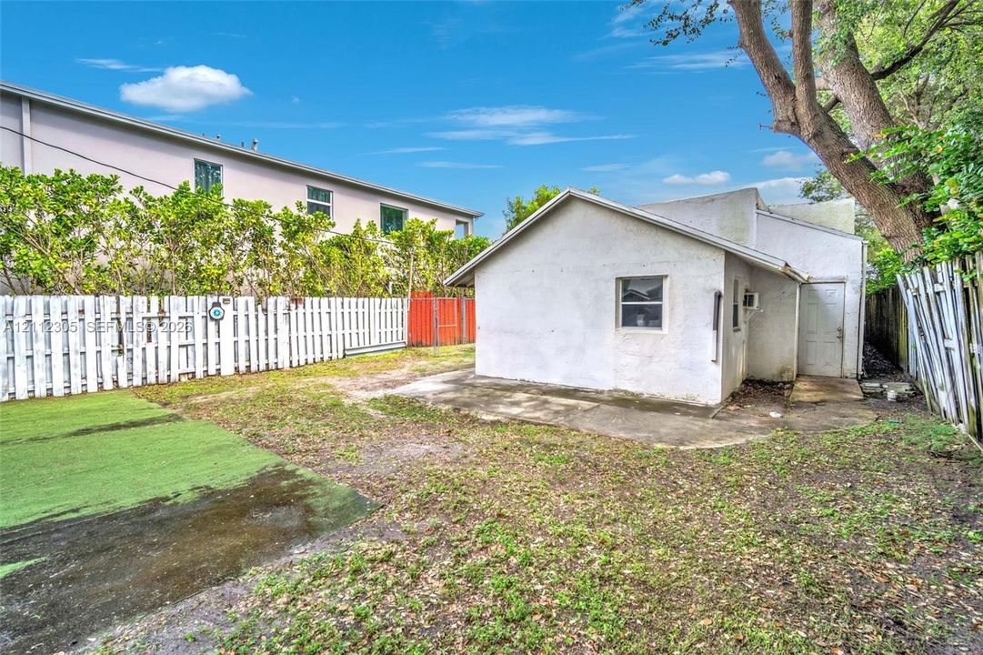 Rear exterior featuring a stucco-finish structure, concrete patio, synthetic turf area, and perimeter privacy fencing