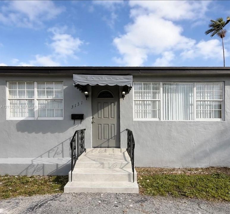 Exterior facade featuring smooth grey stucco, multi-pane windows, arched-top paneled front door, metal scalloped awning, and tiled steps with wrought-iron handrails