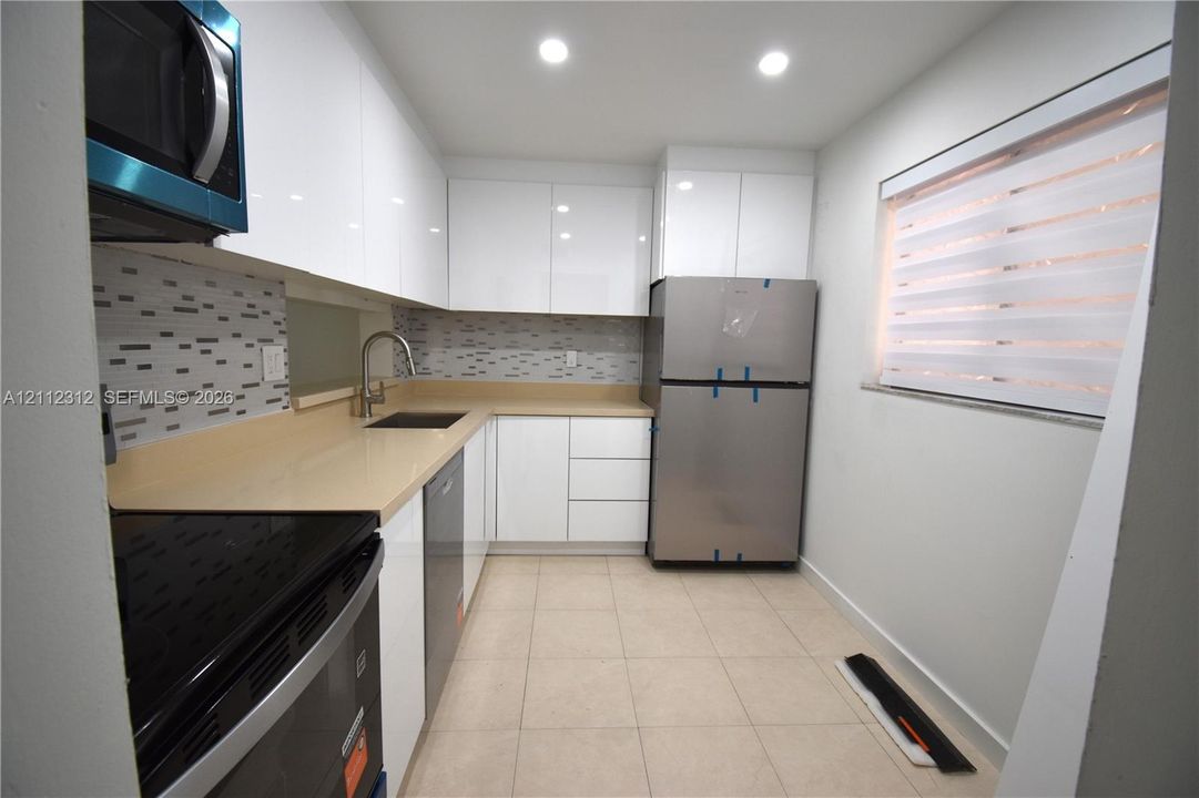 Kitchen featuring high-gloss white cabinetry, solid-surface countertops, mosaic tile backsplash, stainless steel appliances, and ceramic tile flooring