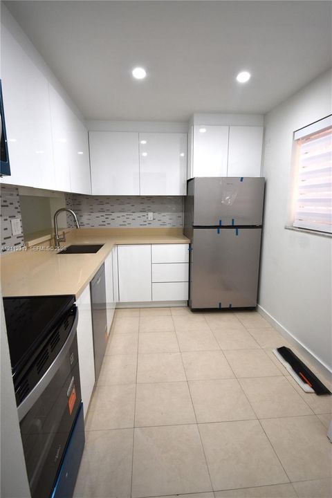 Kitchen featuring high-gloss white cabinetry, solid-surface countertops, a mosaic tile backsplash, stainless steel appliances, and large-format tile flooring