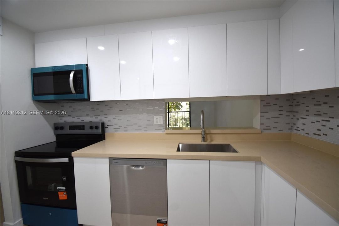 Kitchen featuring high-gloss white cabinetry, neutral solid-surface countertops, stainless steel undermount sink, mosaic tile backsplash, and built-in microwave