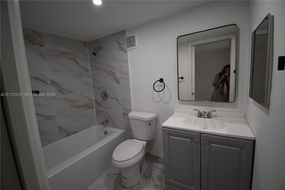 Bathroom featuring a vanity with a white countertop and grey cabinetry, a tub-shower combination with floor-to-ceiling stone-finish tile surround, and a rectangular mirror with rounded corners