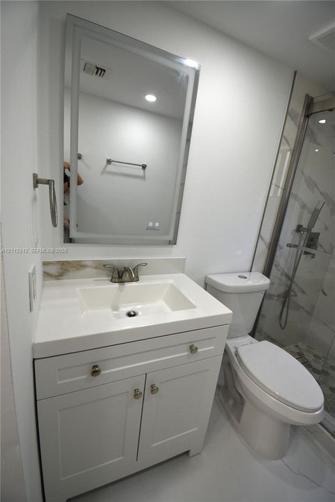 Bathroom vanity with an integrated sink, polished metal faucet, white cabinetry, rectangular LED-lighted mirror, and a walk-in shower with stone-patterned wall tile