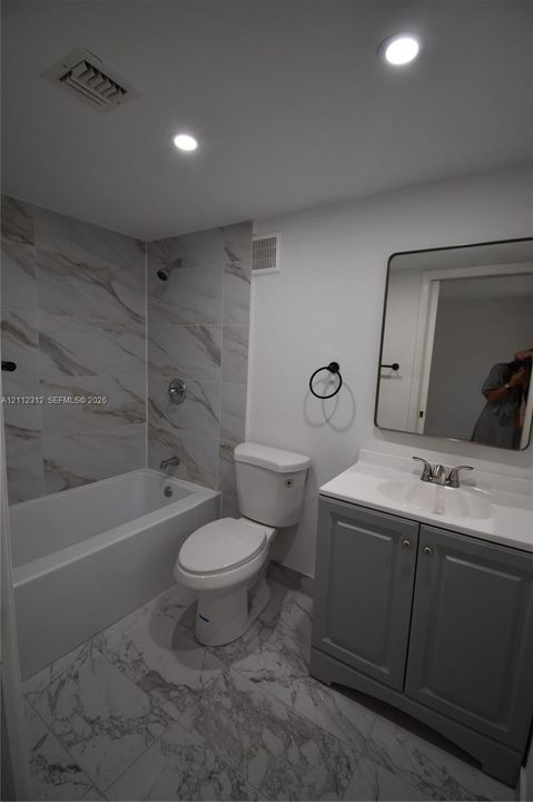 Bathroom featuring a gray vanity cabinet, white countertop with integrated sink, rectangular mirror with rounded edges, tub with large-format tile surround, and tile flooring