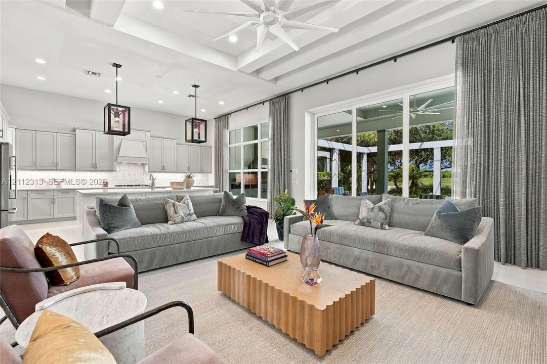 Open-concept living space featuring high coffered ceilings with recessed lighting, oversized sliding glass doors, neutral cabinetry with a stone-finish backsplash, and dual suspended lanterns