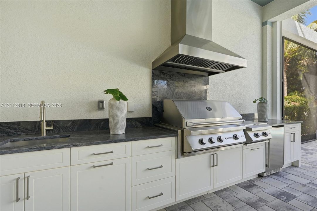 Outdoor kitchen featuring white shaker-style cabinetry, dark natural stone countertops and backsplash, stainless steel built-in grill, commercial-grade ventilation hood, and under-mount sink with modern faucet