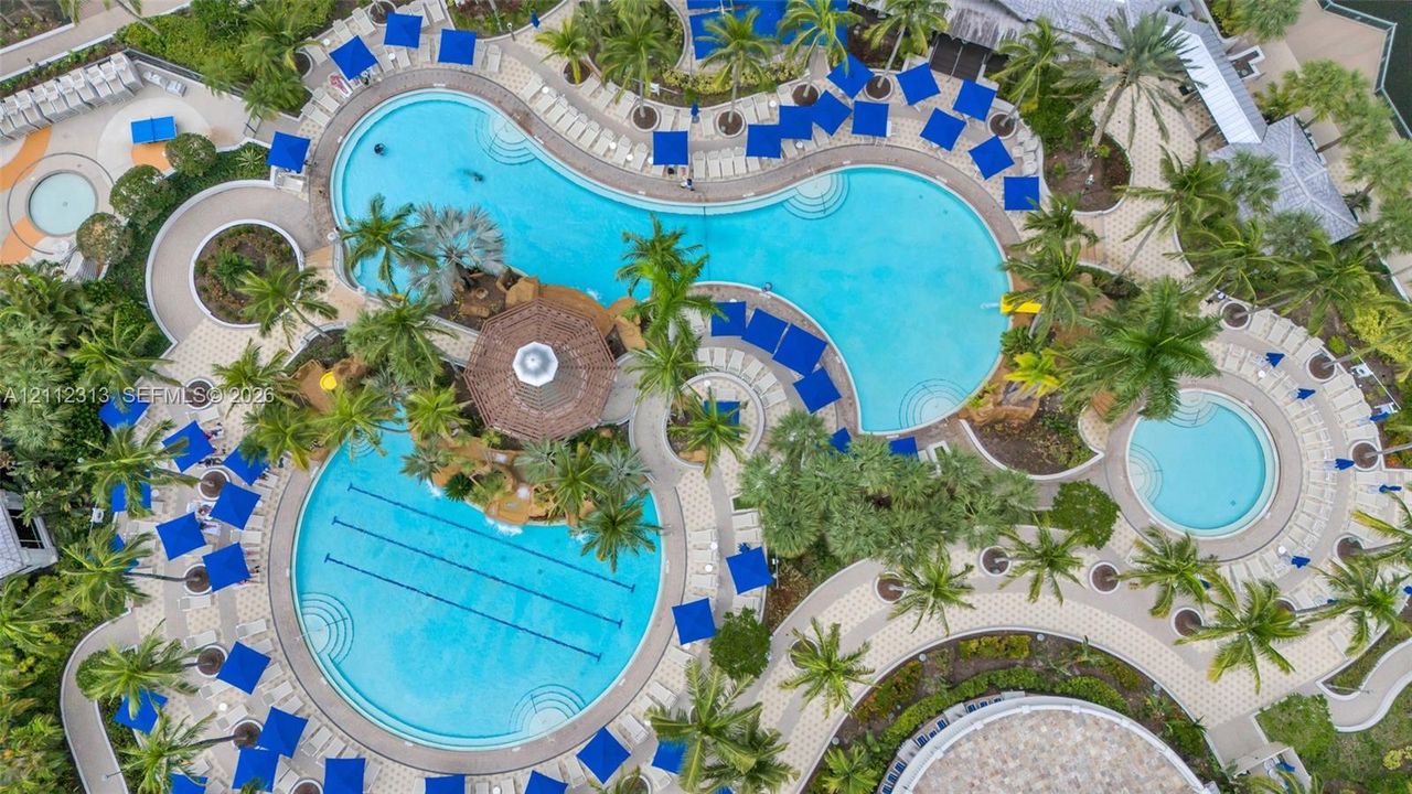 Aerial view of a sprawling pool complex featuring multiple swimming pools, a dedicated lap lane section, a central gazebo, and expansive paved decking surrounded by palm trees
