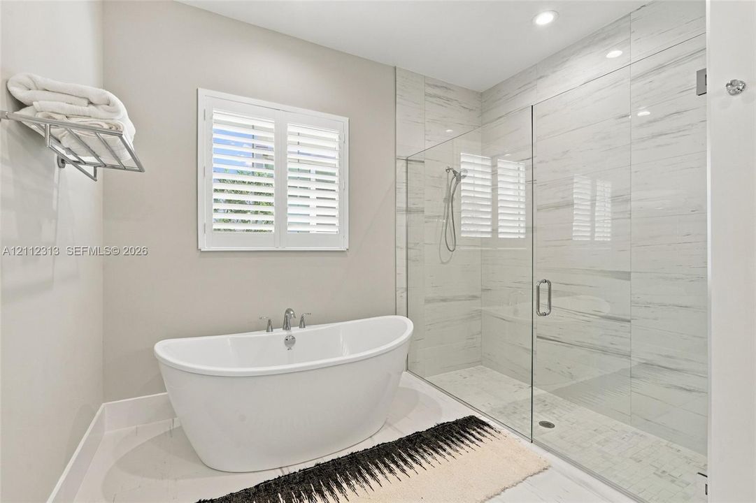 Freestanding soaking tub with chrome fixtures, glass-enclosed walk-in shower with floor-to-ceiling tile, and window shutters