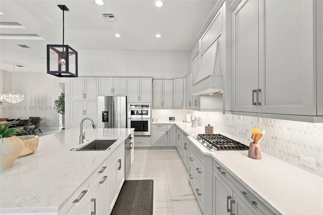 Kitchen featuring light-toned cabinetry, a spacious island with a built-in sink, stone-finish countertops, stainless steel appliances, and a geometric pendant light fixture