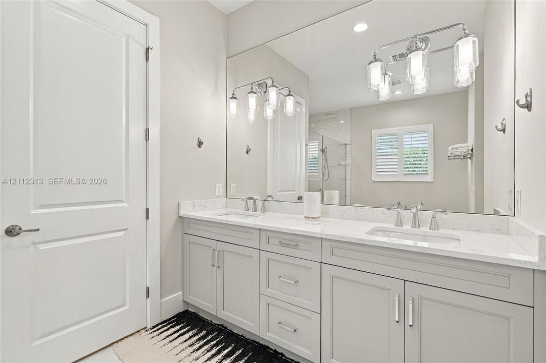 Double vanity featuring light-toned cabinetry, stone-finish countertops, undermount sinks, polished metal fixtures, and a wall-to-wall mirror with multi-bulb integrated lighting