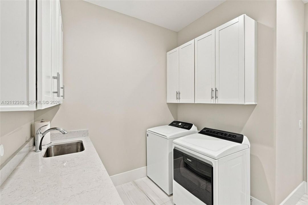 Laundry area featuring white shaker-style wall cabinets, a light-toned countertop, an integrated stainless steel sink with a chrome faucet, and wood-finish flooring