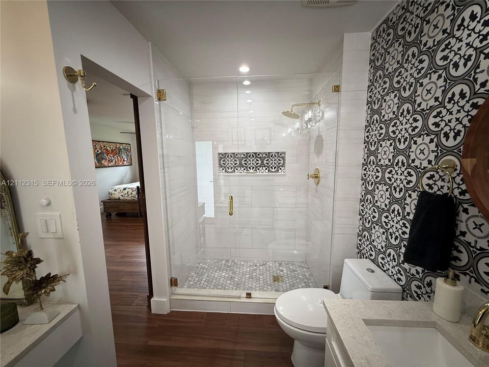 Walk-in shower featuring a glass enclosure, brass-finish fixtures, a decorative tile niche, and penny-round tile flooring, complemented by a patterned tile accent wall and stone-surfaced vanity