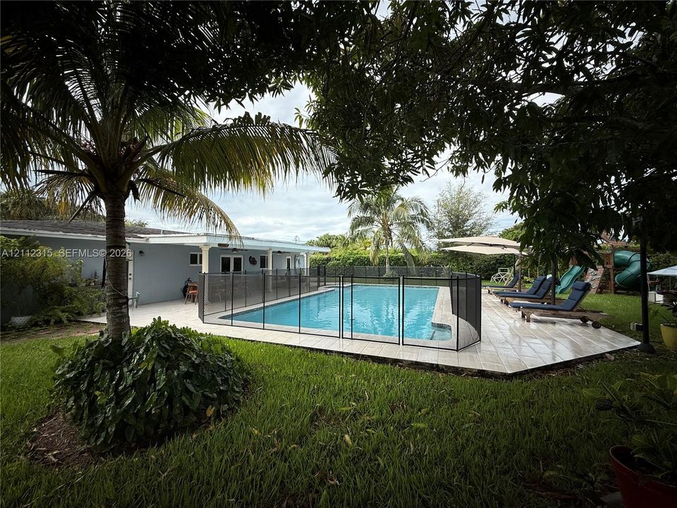 Outdoor living space featuring an in-ground swimming pool, mesh safety fencing, tiled deck, and mature landscaping