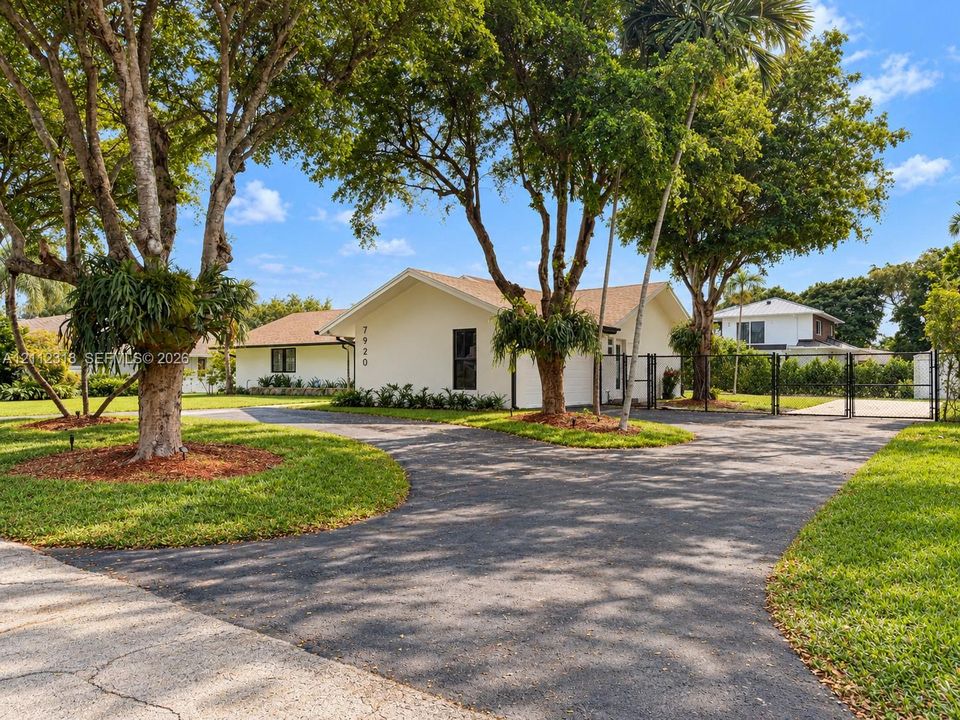 Single-story exterior with light-colored siding, shingle roofing, paved circular driveway, chain-link perimeter fencing, and established mature trees