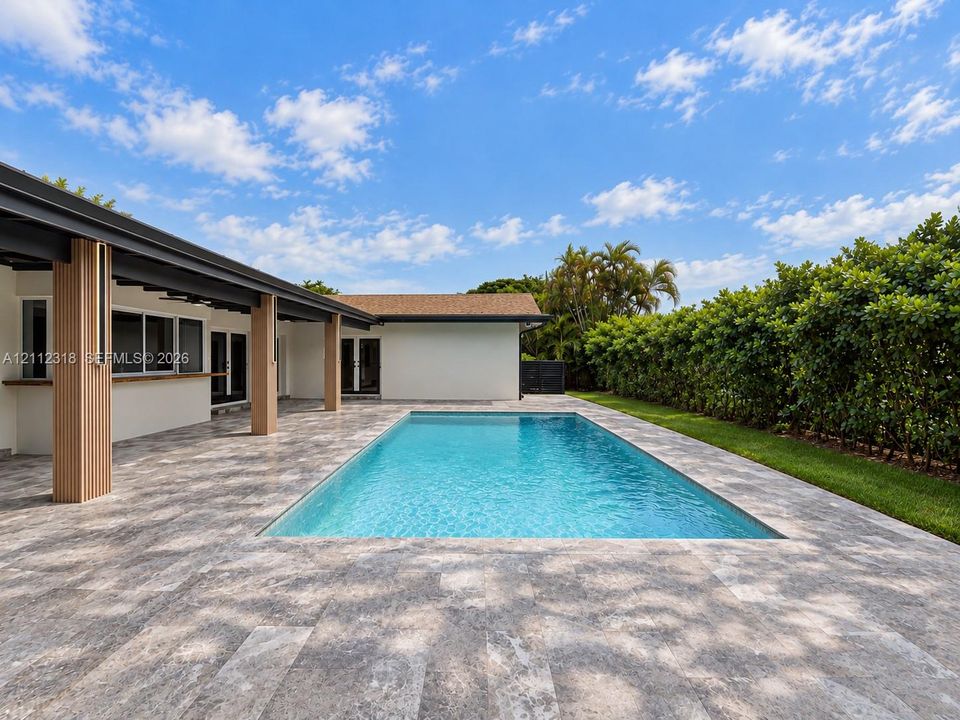 Rectangular swimming pool surrounded by stone-finish paver decking, sheltered covered patio with column detailing, and a manicured hedge perimeter