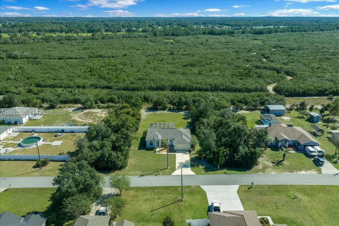 Aerial perspective of a residential property featuring a concrete driveway, a multi-gabled roofline, and an enclosed rear patio area situated on a expansive lot with wooded surroundings