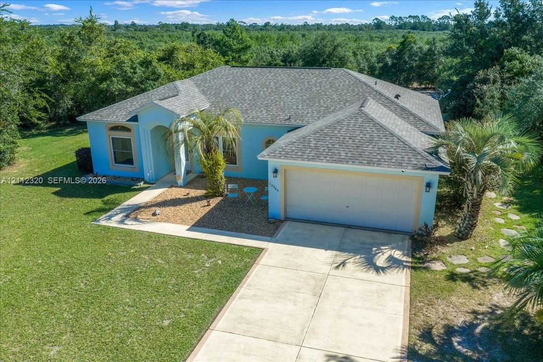 Single-story residence featuring a hipped shingle roof, stucco exterior, two-car garage, concrete driveway, and expansive natural landscaping