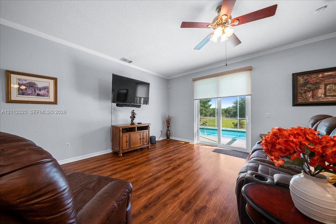 Living area featuring wood-finish flooring, crown molding, ceiling fan with integrated lighting, and sliding glass doors with access to an outdoor pool area
