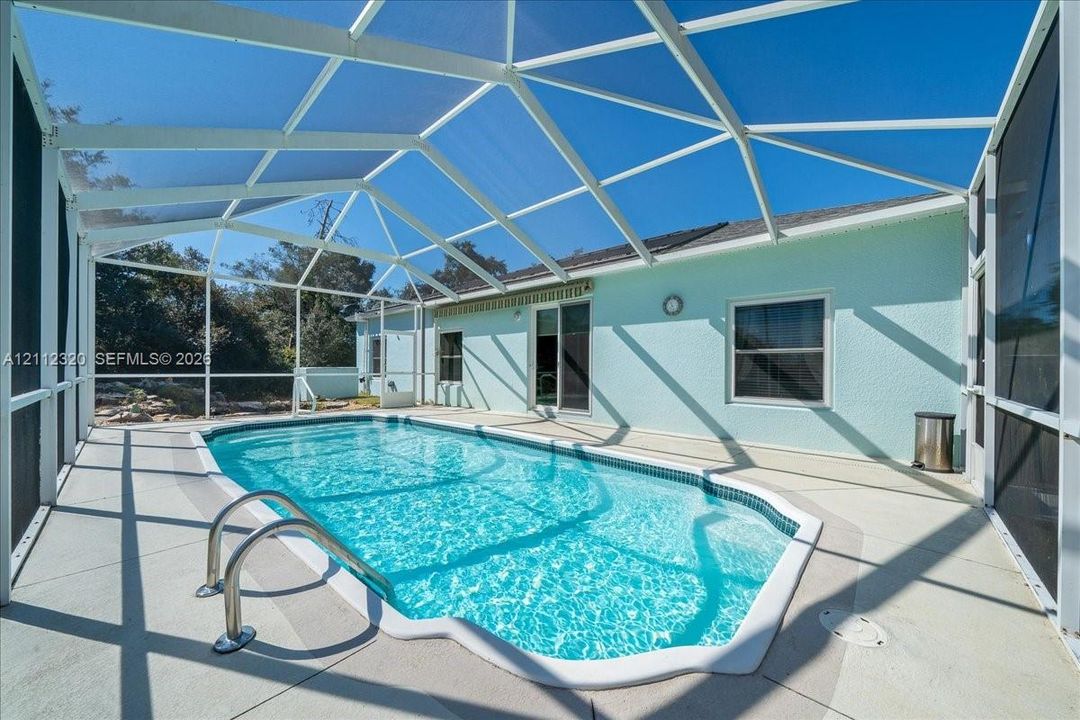 In-ground swimming pool enclosed by a white-frame screen cage, featuring concrete decking and a textured exterior finish