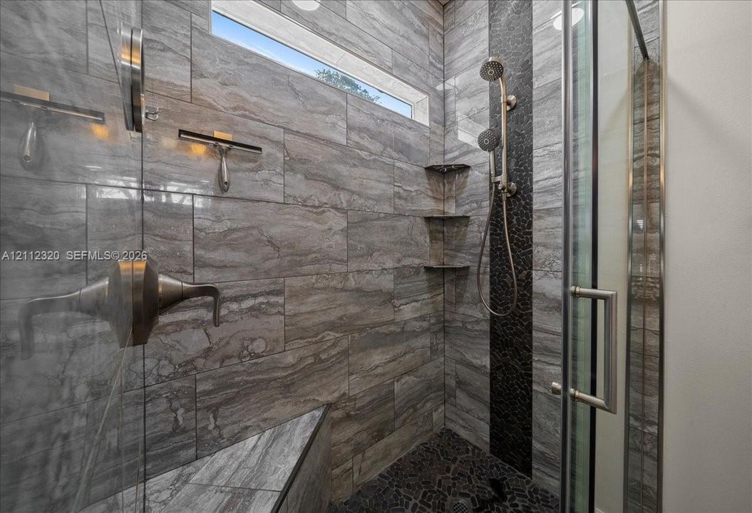 Walk-in shower featuring large-format stone-tile walls, a built-in corner bench, a mosaic-tiled accent wall, a handheld shower fixture, and a transom window