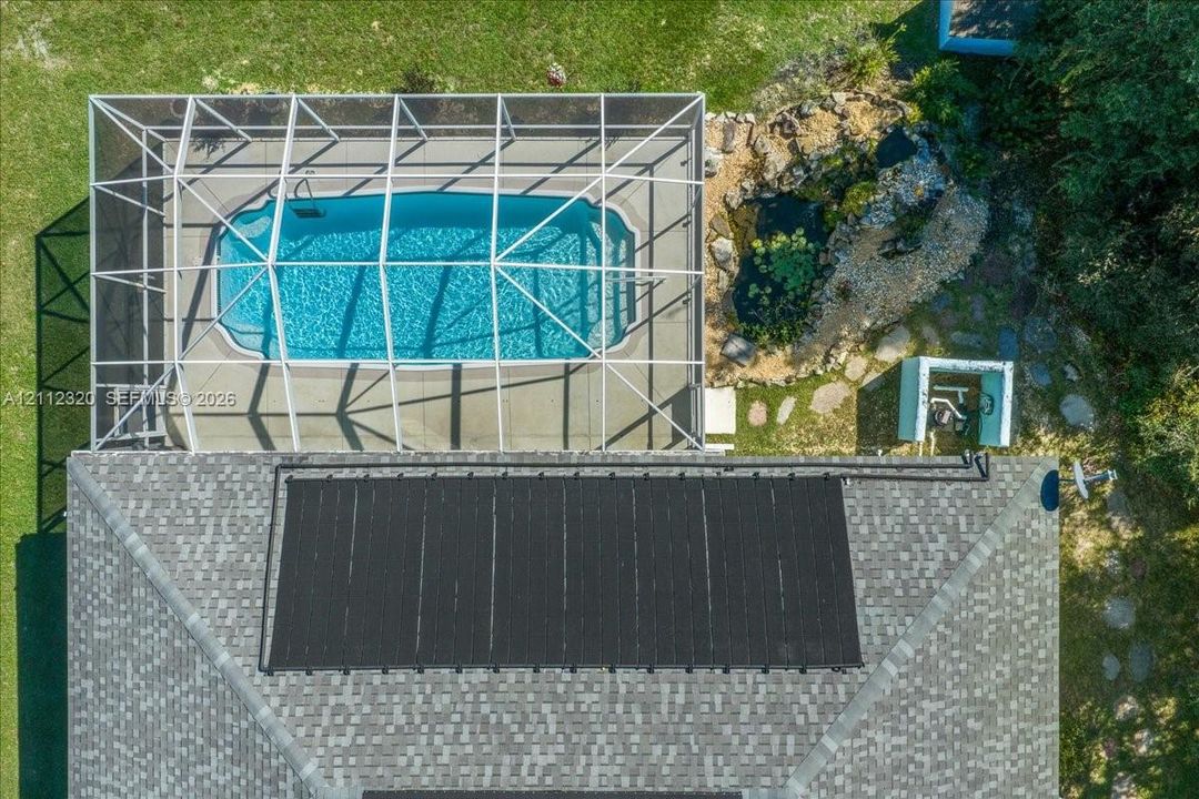 Screen-enclosed swimming pool featuring a concrete deck, roof-mounted solar heating panels, and a rock-lined garden pond