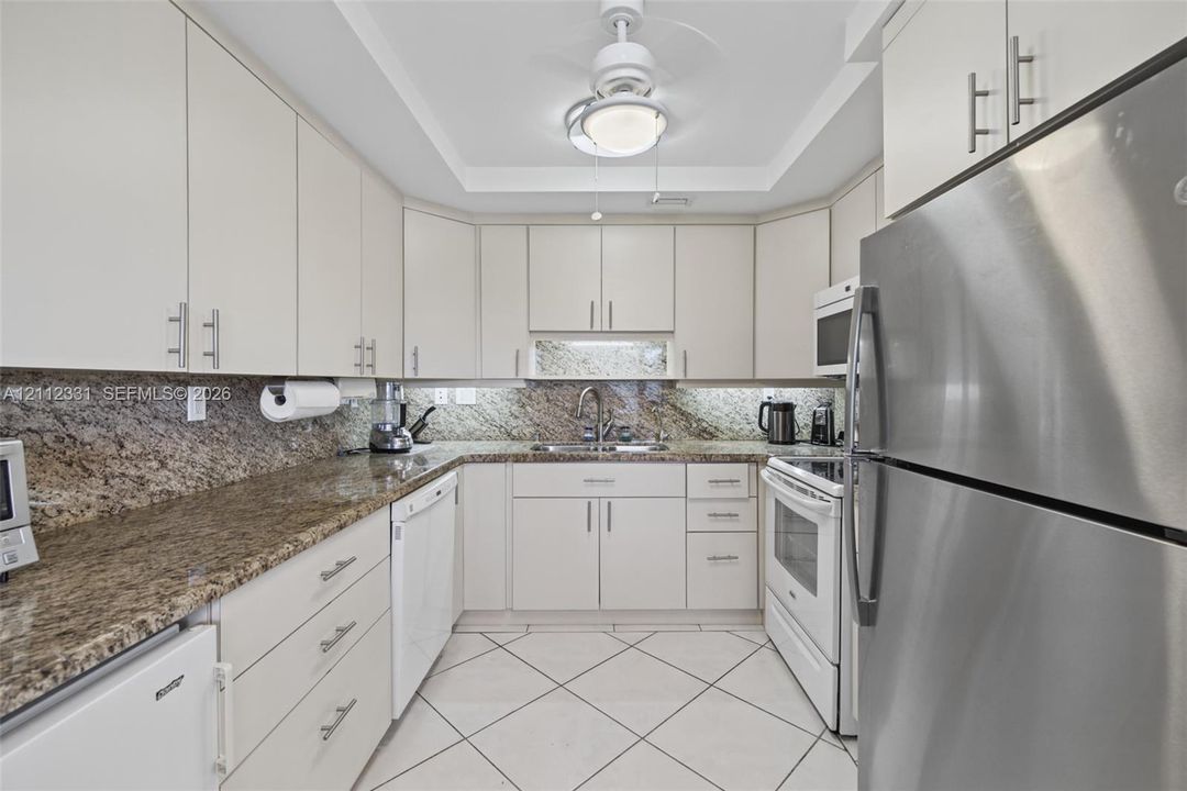 Kitchen featuring light-toned cabinetry with brushed metal pulls, stone-composite countertops, stainless steel refrigerator, white appliances, and diagonal-set tile flooring