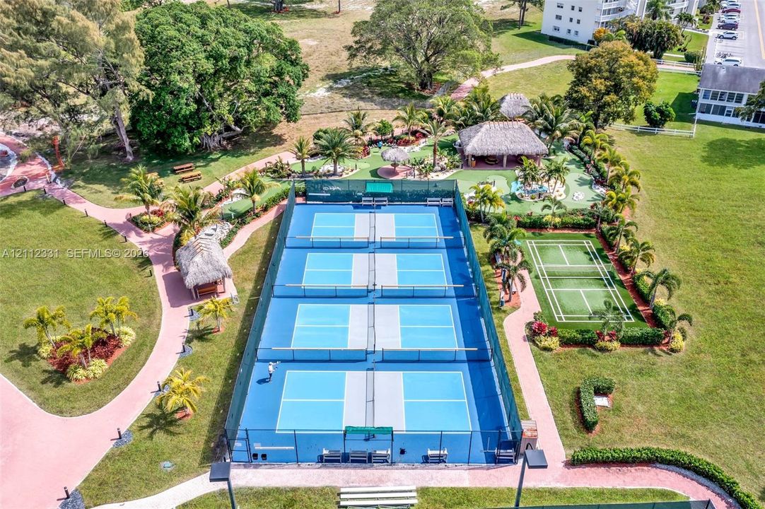 Recreational complex featuring four pickleball courts, a grass tennis court, multiple thatch-roofed pavilions, and landscaped walking paths