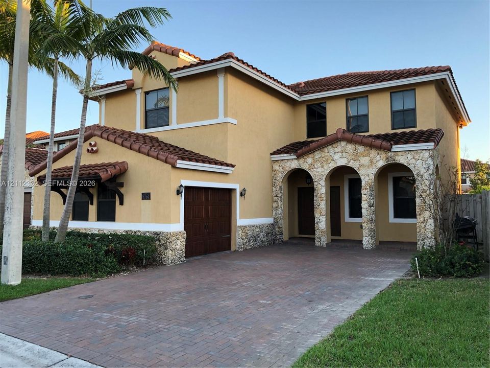 Two-story Mediterranean-style exterior featuring stucco walls, decorative stone masonry, a multi-gabled terracotta tile roof, an arched covered entryway, and a paver driveway