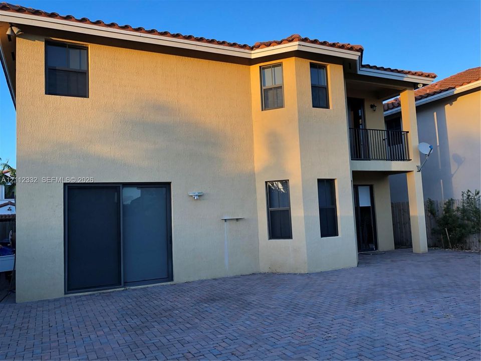 Stucco exterior featuring a clay tile roof, a covered second-story balcony with a metal railing, and a paved brick patio
