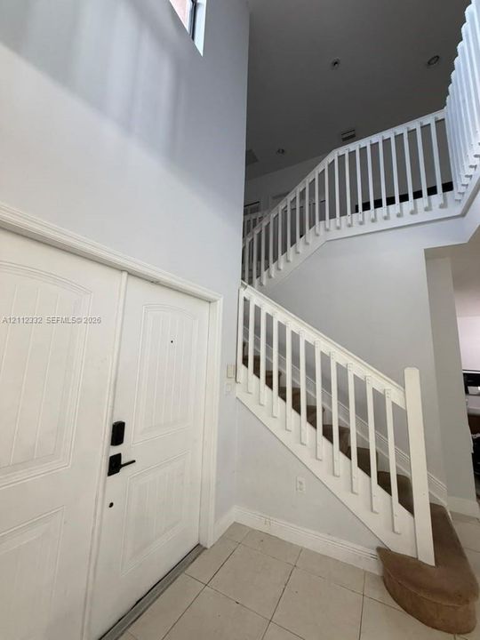 Double-height entry featuring white double doors, neutral tile flooring, and a staircase with a white spindle railing