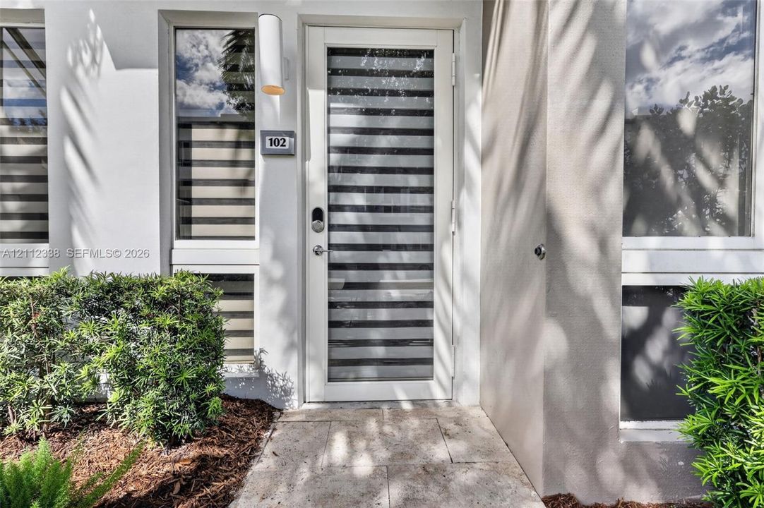 Exterior entry featuring a white door with horizontal glass panes, stone-finish paving, modern cylindrical wall-mounted lighting, and manicured shrubbery