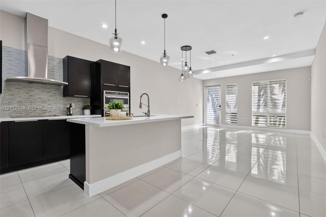 Kitchen featuring dark cabinetry, quartz countertops, a stone-tiled backsplash, stainless steel range hood, and polished tile flooring