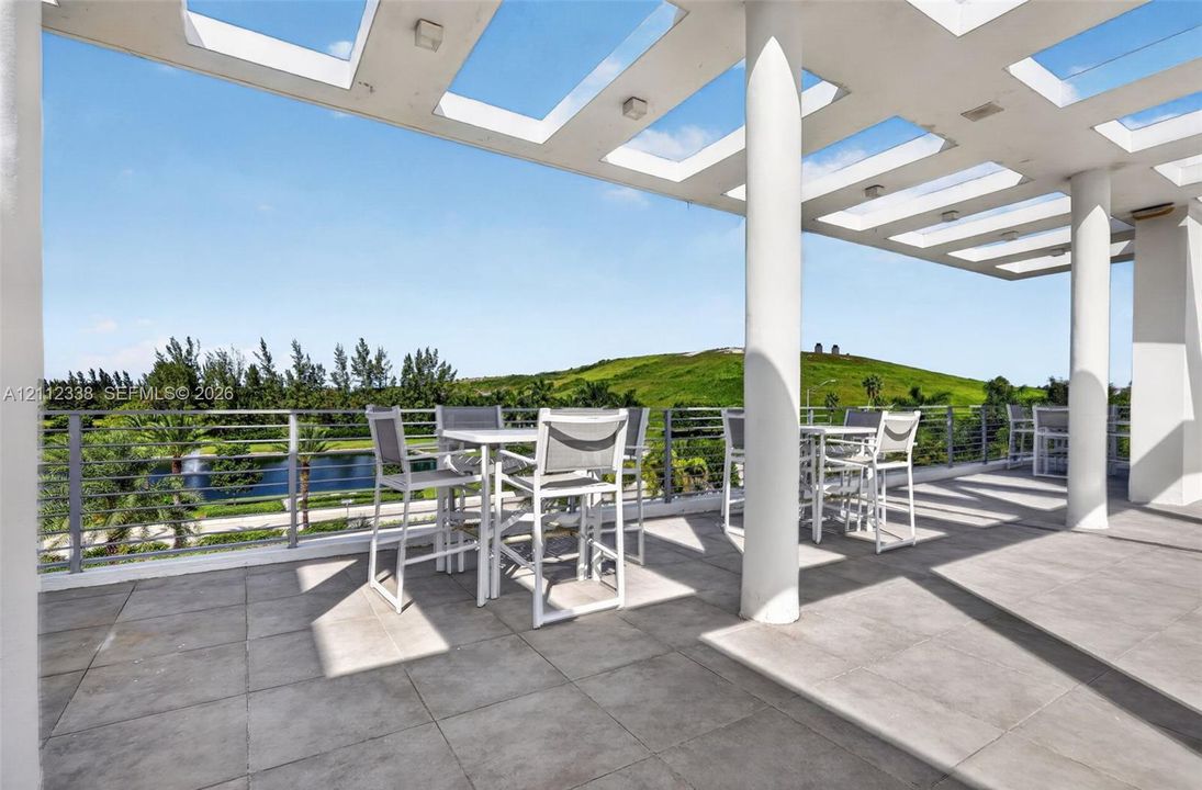 Covered terrace featuring a pergola-style ceiling, structural support columns, oversized tile flooring, and a metal cable railing system overlooking a landscape backdrop