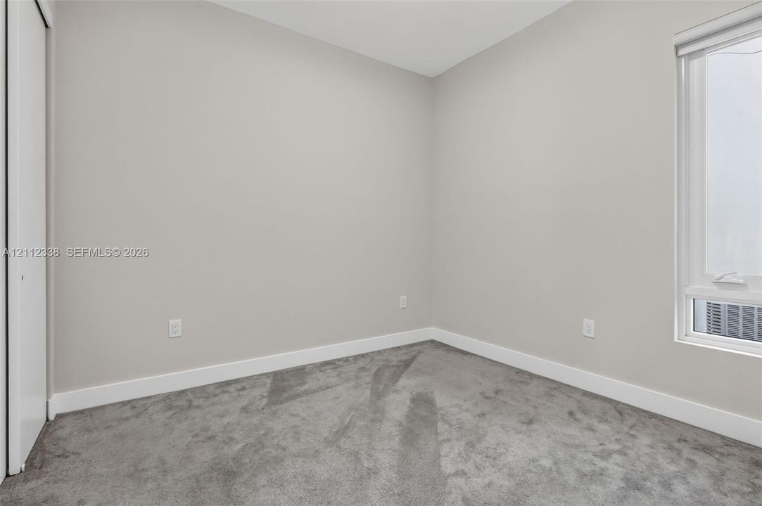 Neutral-toned interior space featuring soft wall-to-wall carpeting, crisp white walls, and a window with an integrated roller shade