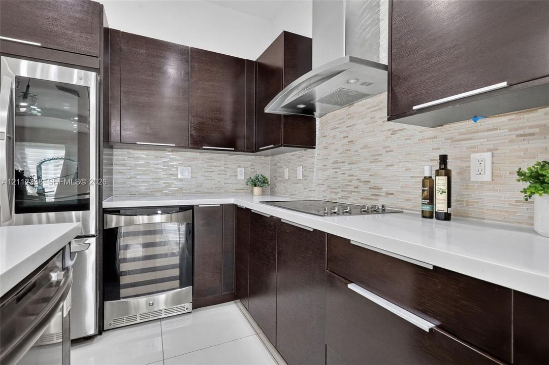 Dark wood cabinetry with modern hardware, white quartz countertops, stainless steel appliances, tiled backsplash, and light-tone tile flooring