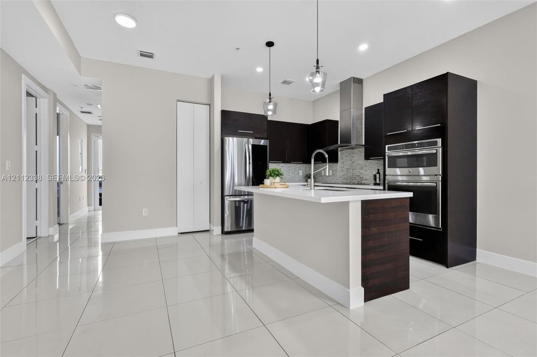 Kitchen featuring dark wood-finish cabinetry, a central island with white countertop, stainless steel double wall ovens, a stainless steel range hood, and tile flooring