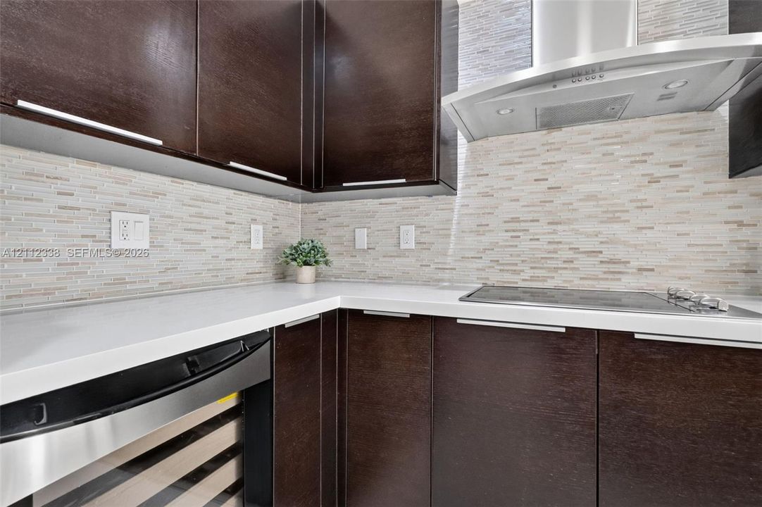 Dark wood-finish cabinetry featuring sleek hardware, white solid-surface countertops, horizontal linear tile backsplash, stainless steel range hood, and integrated under-counter beverage chiller