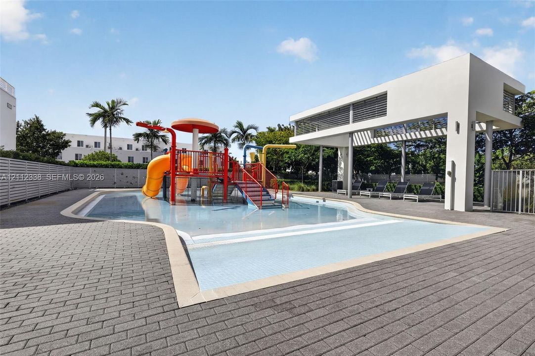 Splash pad area featuring integrated water slides, a central mushroom-shaped fountain, a concrete paver deck, and a modern open-air covered pavilion