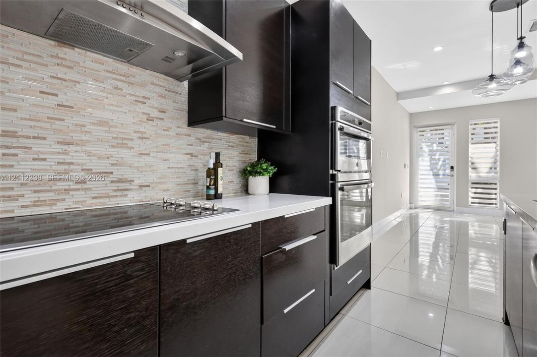 Kitchen featuring dark-finish flat-panel cabinetry, light-toned stone countertops, a linear mosaic tile backsplash, stainless steel built-in double ovens, and large-format porcelain tile flooring
