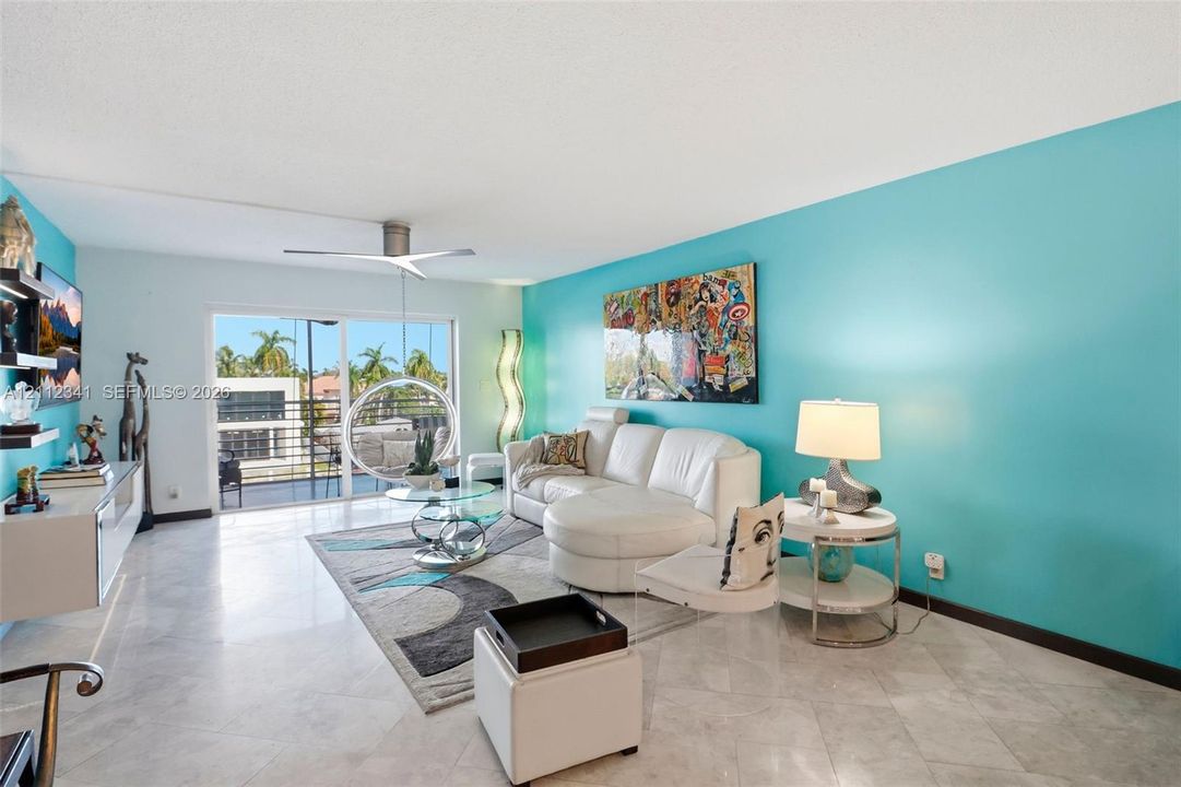 Open-concept living area featuring light-toned stone tile flooring, a multi-blade ceiling fan, built-in wall-mounted cabinetry, and large sliding glass doors providing access to a private balcony