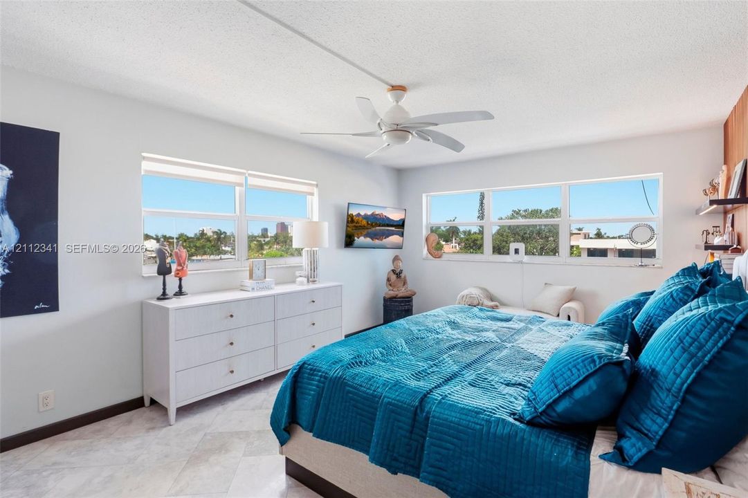 Bedroom featuring stone-finish flooring, multi-pane windows with exterior scenery, a white ceiling fan, and wood-finish wall paneling