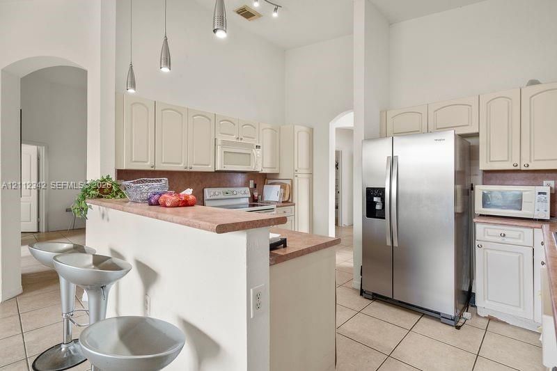 Kitchen featuring high ceilings, off-white cabinetry, solid-surface countertops with a breakfast bar, ceramic tile flooring, and stainless steel side-by-side refrigerator