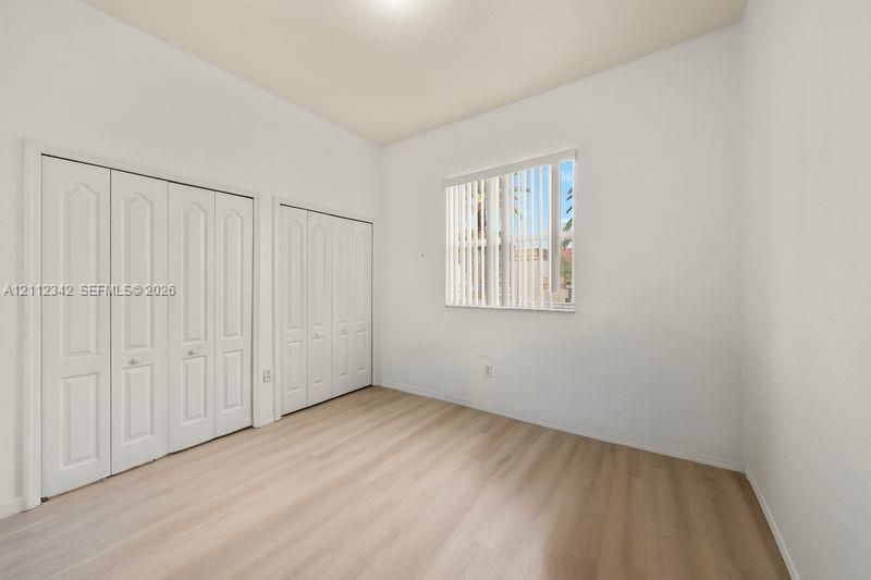 Interior space featuring light-toned wood-finish flooring, dual sets of white paneled bifold closet doors, and a window with vertical blinds