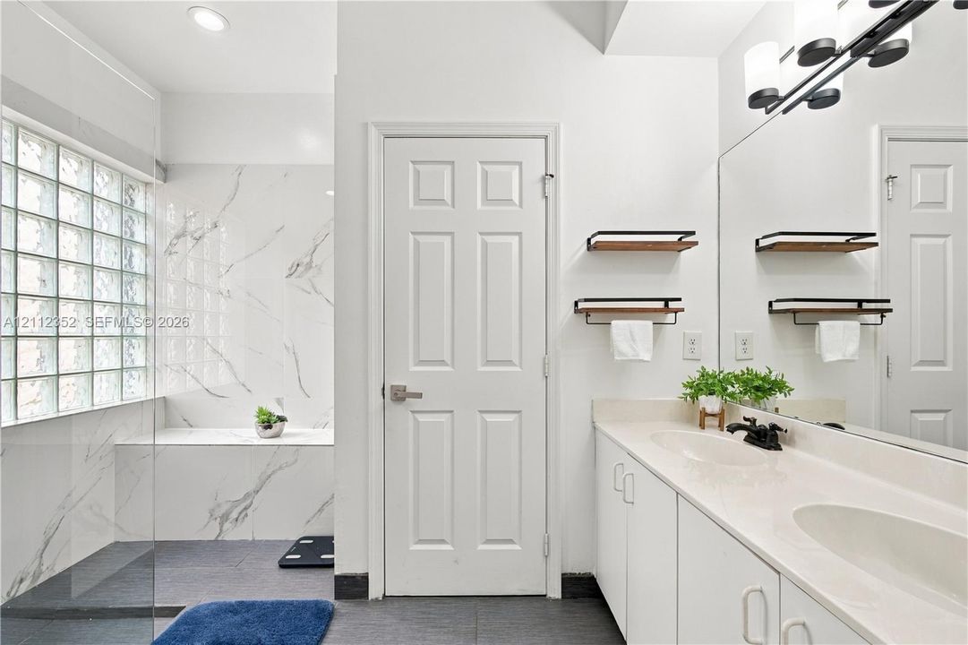 Bathroom featuring a walk-in shower with stone-finish surround, glass block window, double vanity with solid surface countertop, and matte black fixtures